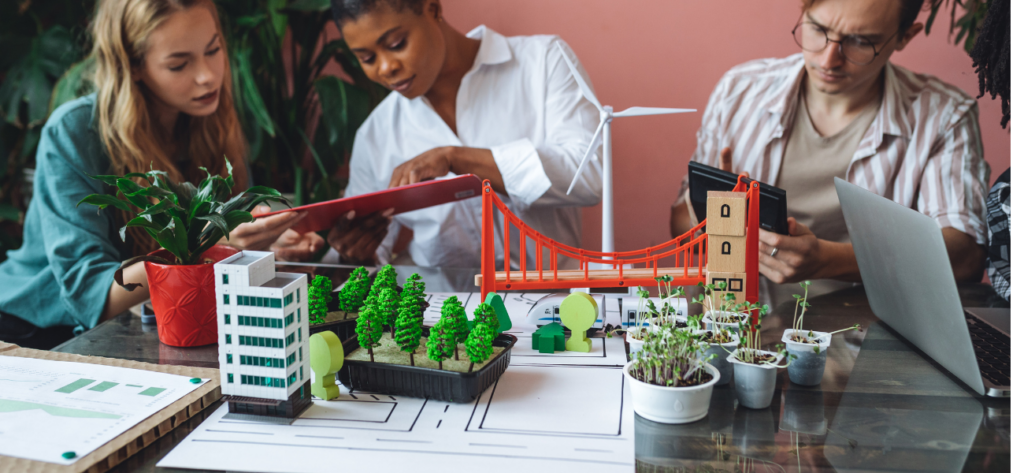 Trois personnes, des architectes, se tiennent derrière des maquettes d'éléments constitutifs d'un projet urbain : pont, arbre...